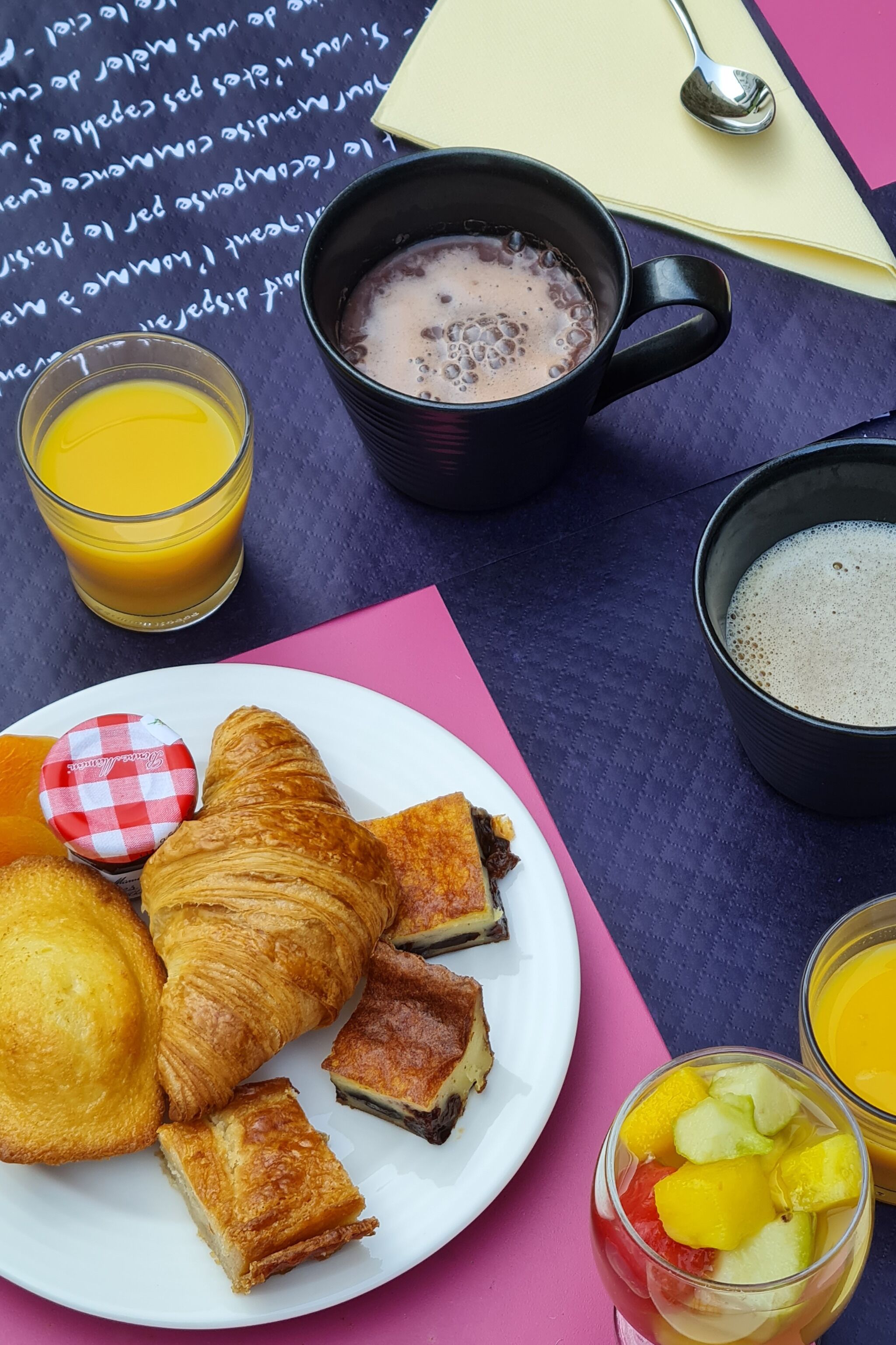 Une table mise pour le petit-déjeuner avec des croissants, une assiette de fruits, une tasse de café et un verre de jus d’orange.