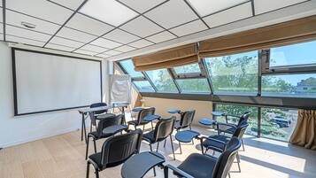 A conference room with a projector screen, multiple tables, chairs, and large windows offering a view of the parking lot and trees.
