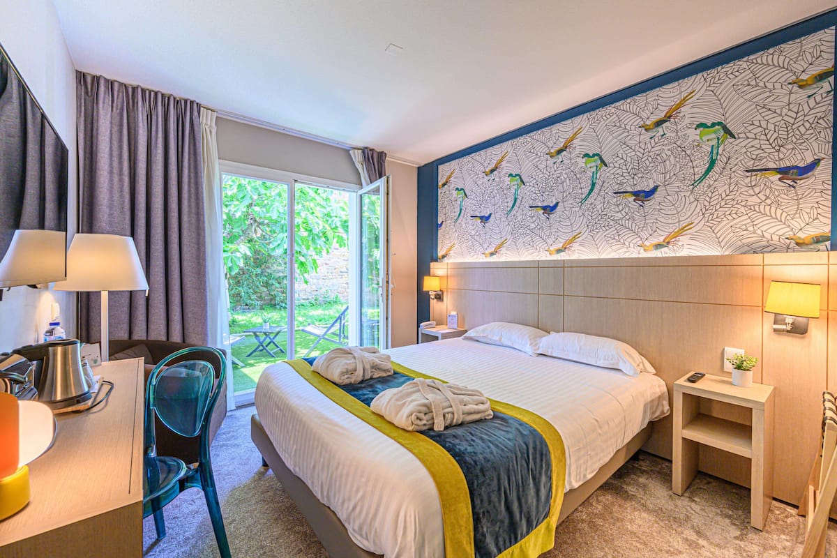 A hotel room with a large bed, a desk with a lamp, a chair, a nightstand with a lamp, and a mural of birds on the wall.