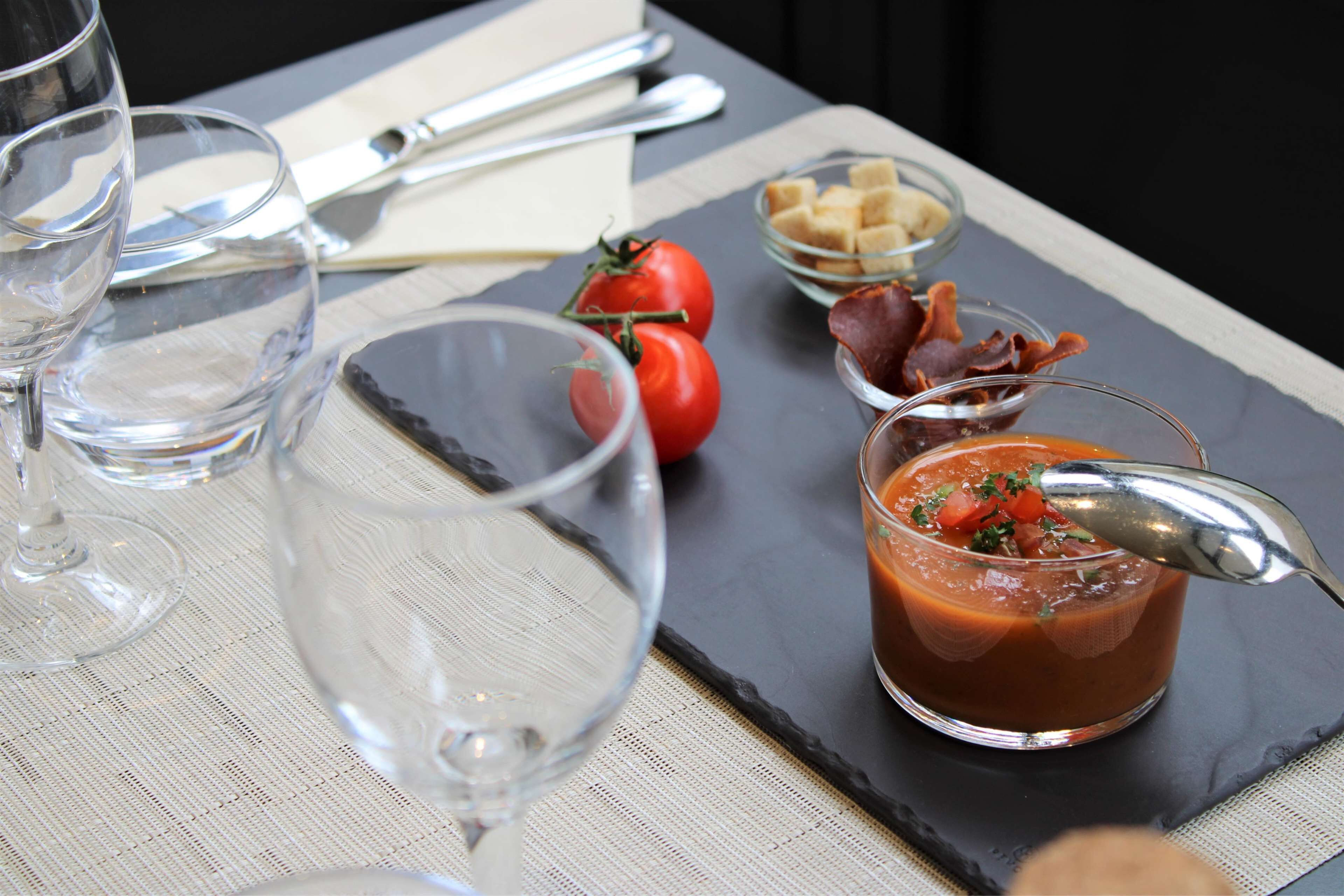 A table with a glass of water, a glass of tomato soup garnished with herbs, a bowl of bread cubes, and a small bowl of bacon.