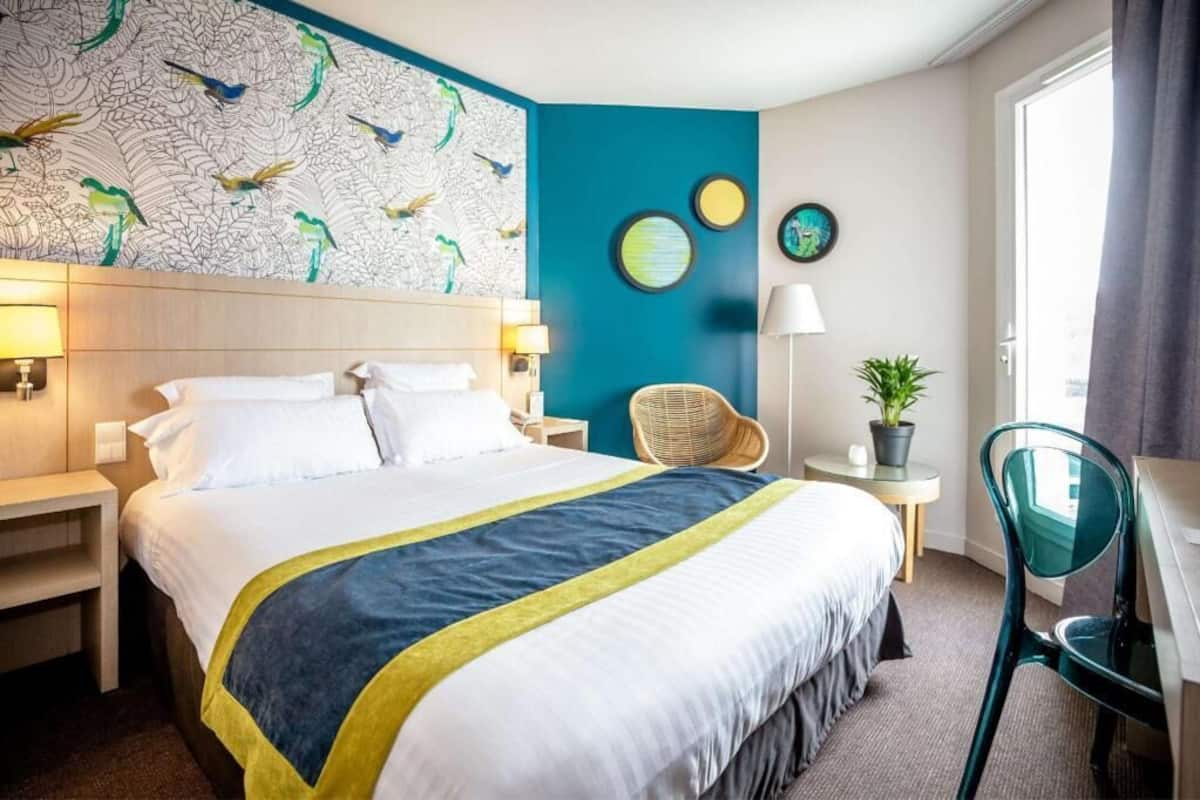A hotel room with a large bed, a desk, a chair, a wall with a bird mural, and a door.