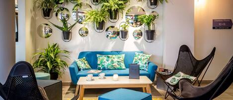 Un vestíbulo moderno con un sofá azul, sillas negras, una mesa de centro y una variedad de plantas en macetas dispuestas en las paredes.
