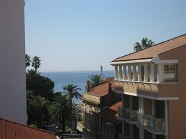 A coastal building with a balcony overlooking the sea, palm trees, and a lighthouse in the distance.
