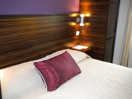 A neatly made bed with a white mattress and a single maroon pillow, positioned against a wooden headboard with built-in lighting.