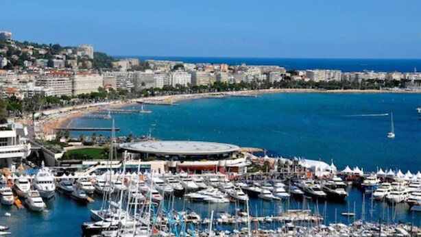 Un puerto deportivo con numerosas embarcaciones amarradas, un paisaje urbano costero con edificios y un cielo azul despejado.