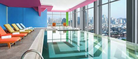 A modern indoor swimming pool with a city view, colorful lounge chairs, and a glass barrier.