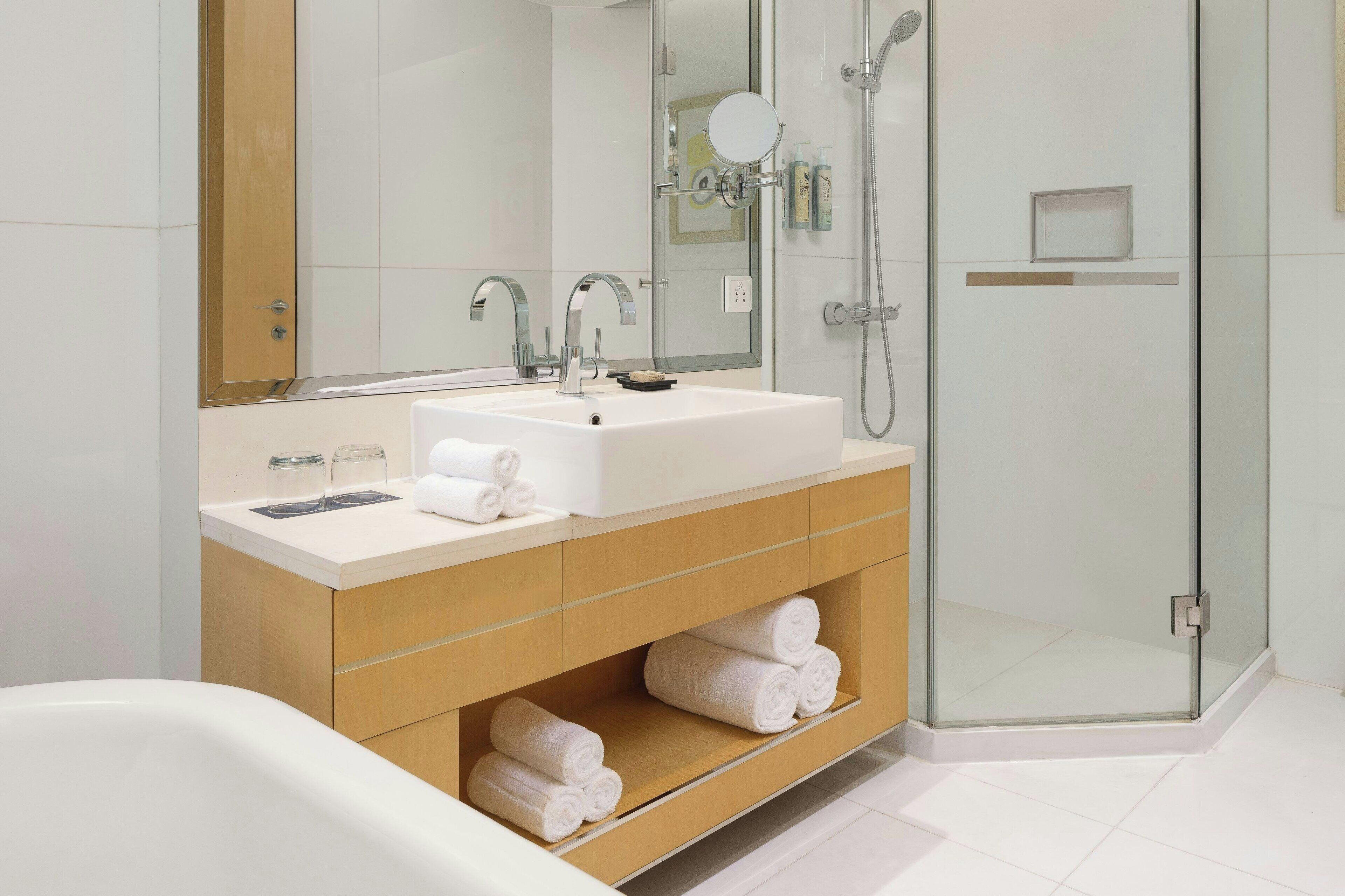Un baño moderno con un lavamanos grande, dos espejos, cabina de ducha con mampara de vidrio y toallas ordenadas en estantes.