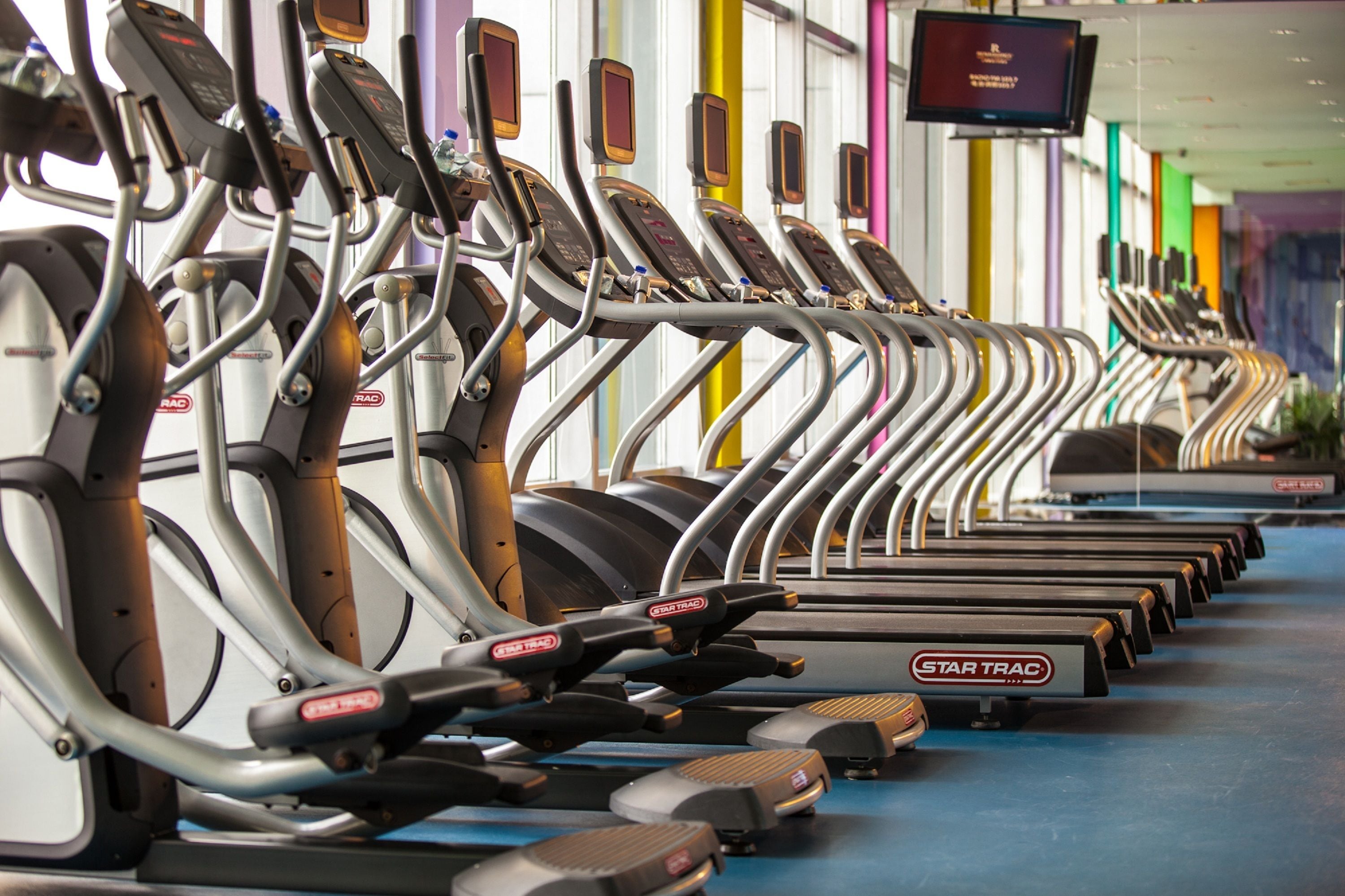 En rad löpband av märket Star Trac i ett gym, med en tv-skärm ovanför dem.