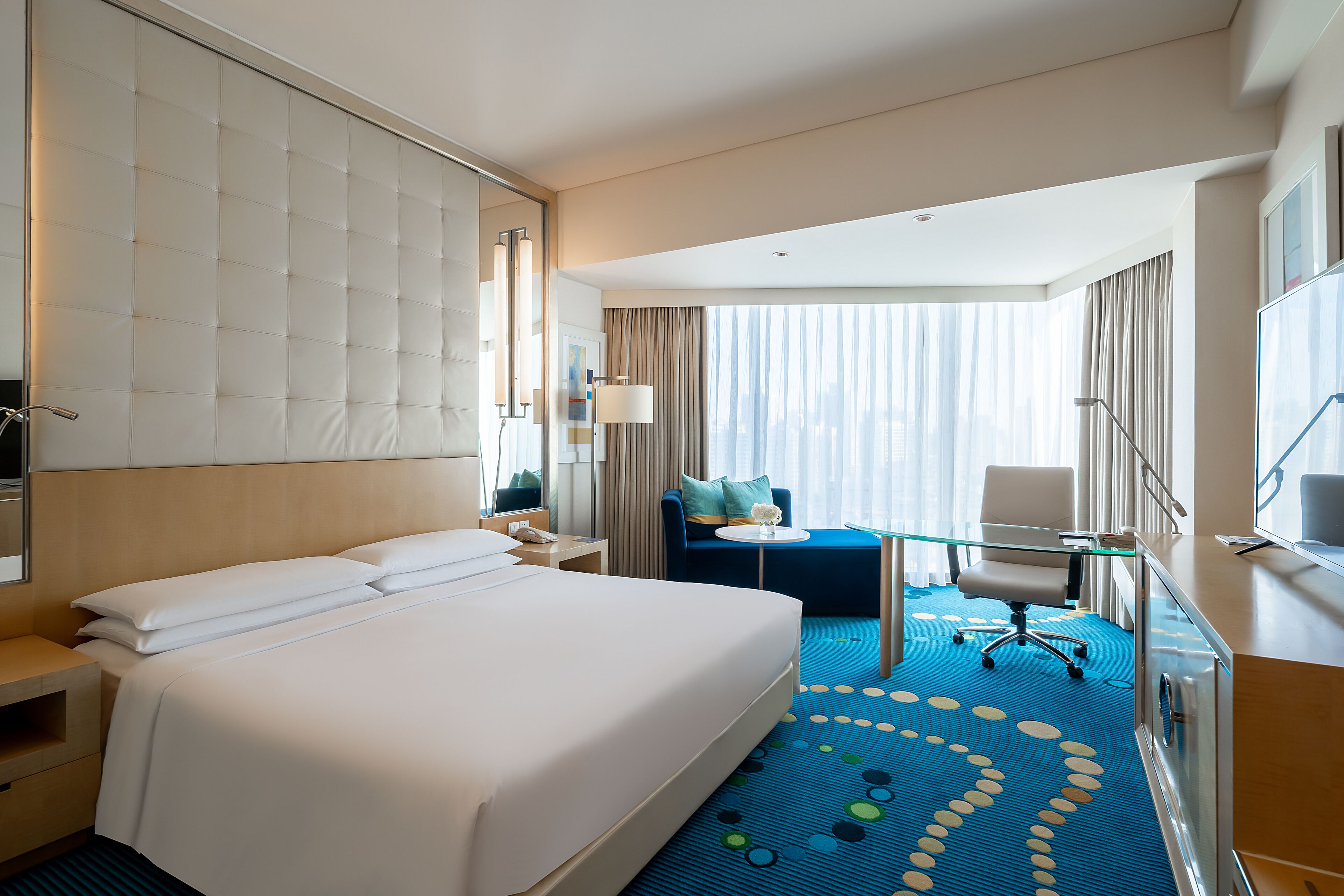 A hotel room with a large bed, a desk with a chair, a small table, and a large window with curtains.
