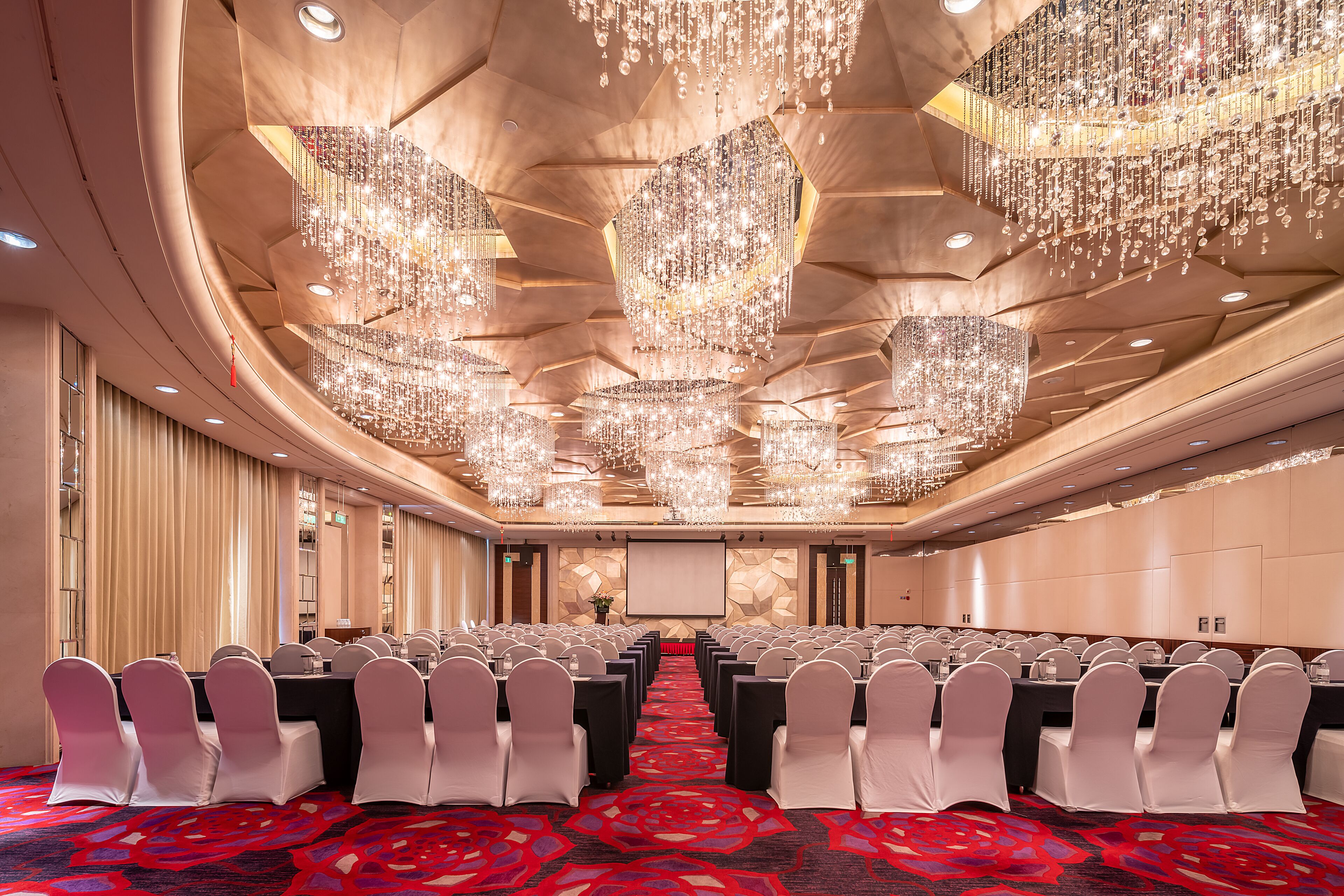 A conference room with rows of tables and chairs, a large screen, and a ceiling with multiple chandeliers.