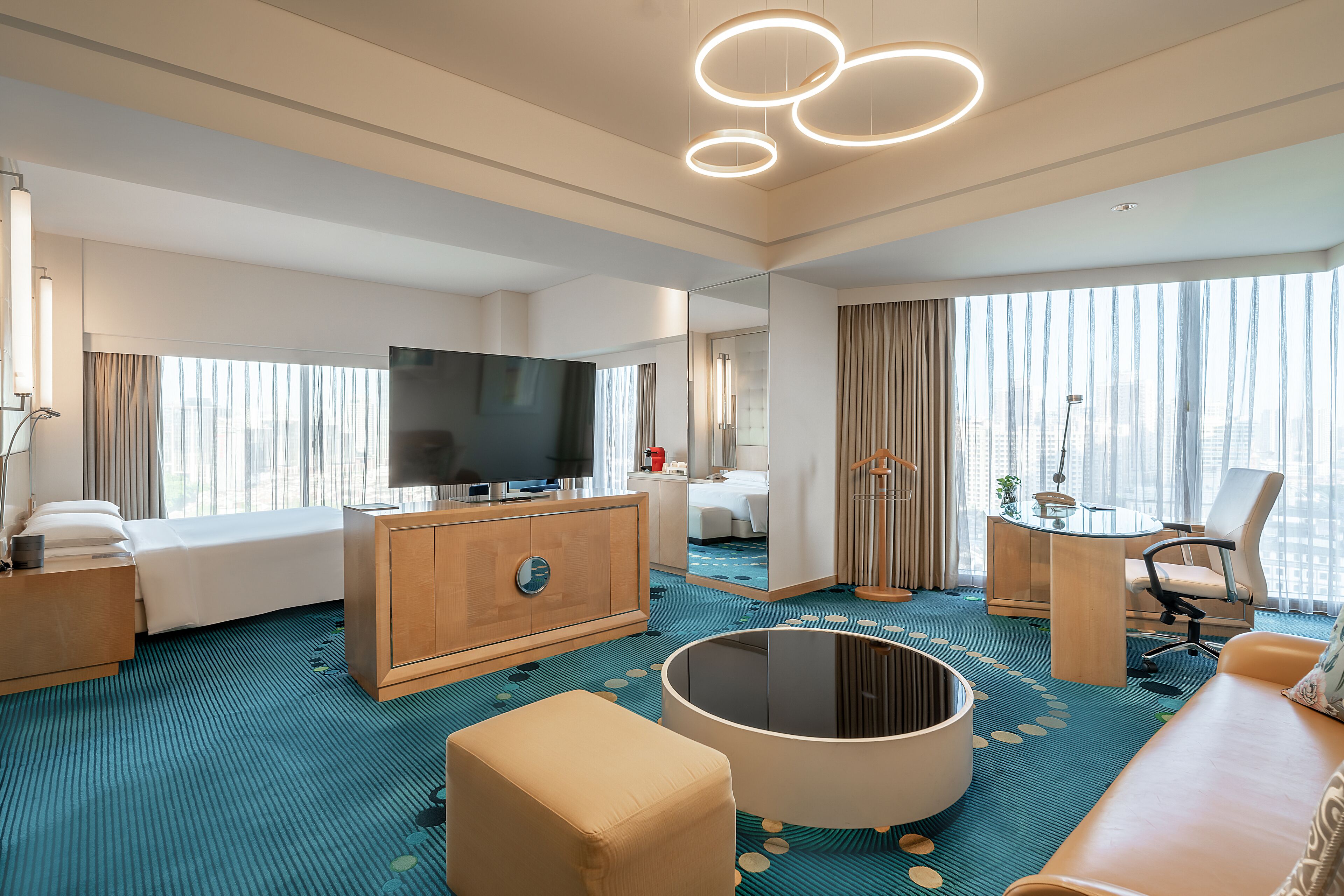 A modern hotel room with a large bed, a TV, a round coffee table, a sofa, and a desk with a chair.