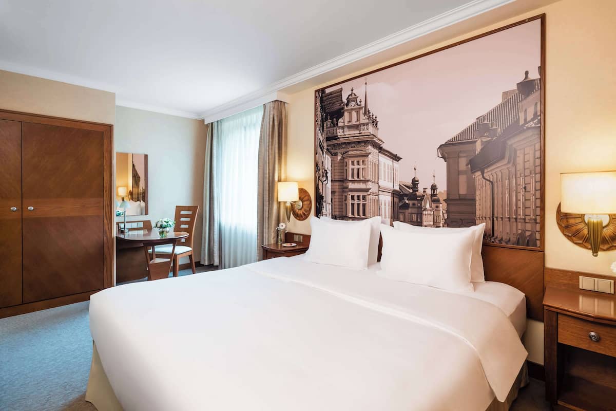Habitación de hotel con una cama grande, un escritorio con silla, una mesita de noche con lámpara y una lámpara de pared.