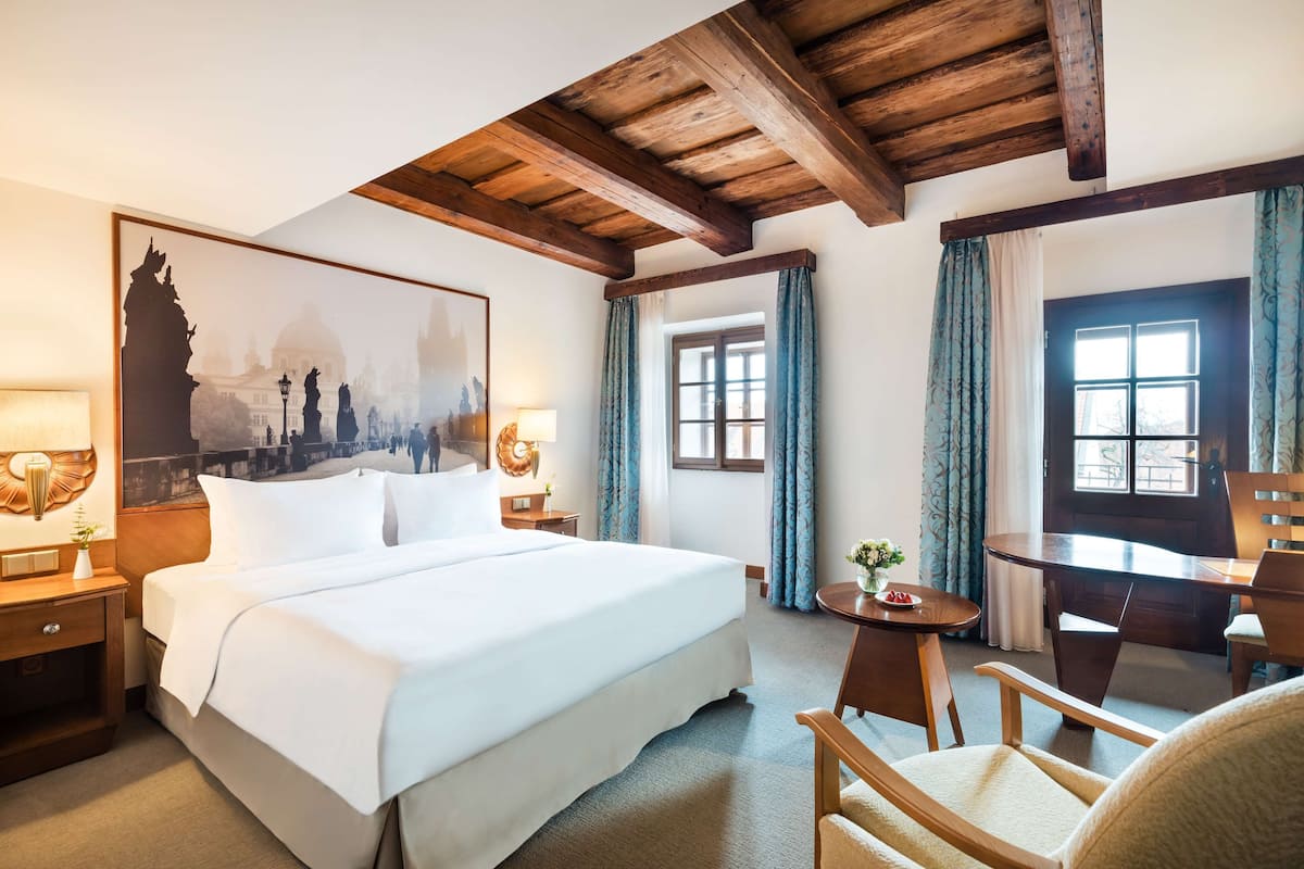 Habitación de hotel con una cama grande, un escritorio y una silla. Se ve una ventana con cortinas y un cuadro en la pared.