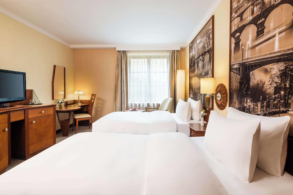 Habitación de hotel con dos camas, un escritorio con televisor y un ventanal con cortinas.