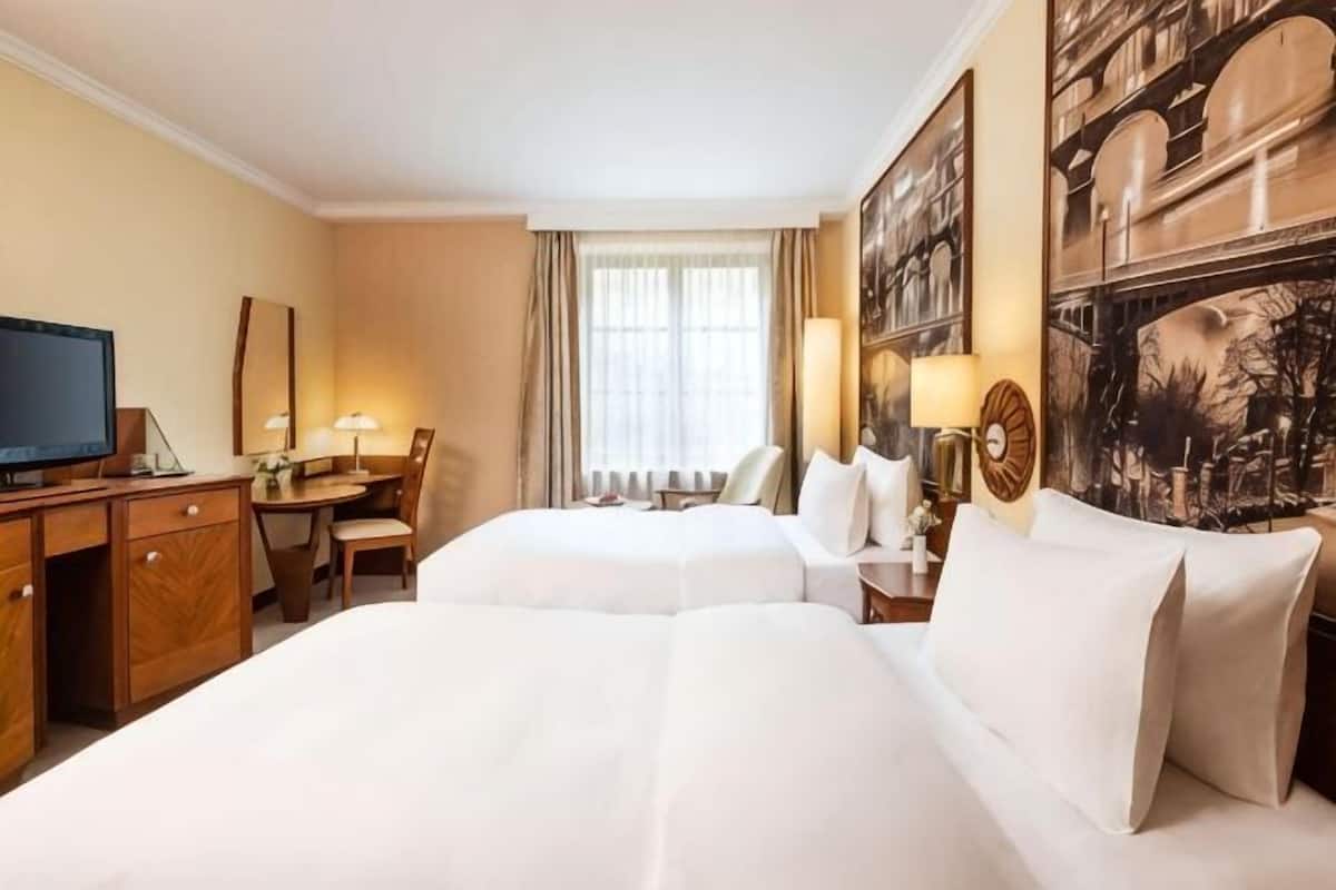 Habitación de hotel con dos camas, un escritorio, televisión y un ventanal con cortinas.