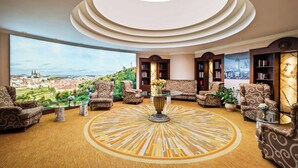 Un salon spacieux avec une table basse au centre, entouré de canapés et de fauteuils, une grande fenêtre donnant sur la ville et un tapis rond à motifs.