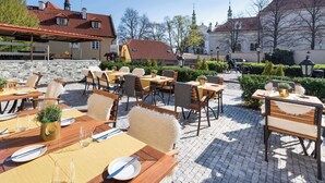Un espace extérieur aménagé pour les repas, avec des tables et des chaises, un mur en pierre et des bâtiments en arrière-plan.
