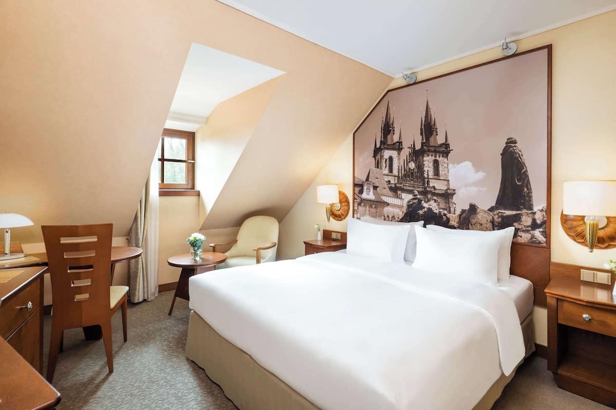 Habitación de hotel con una cama grande, un escritorio con una silla, una mesita con un florero y un cuadro de un castillo en la pared.