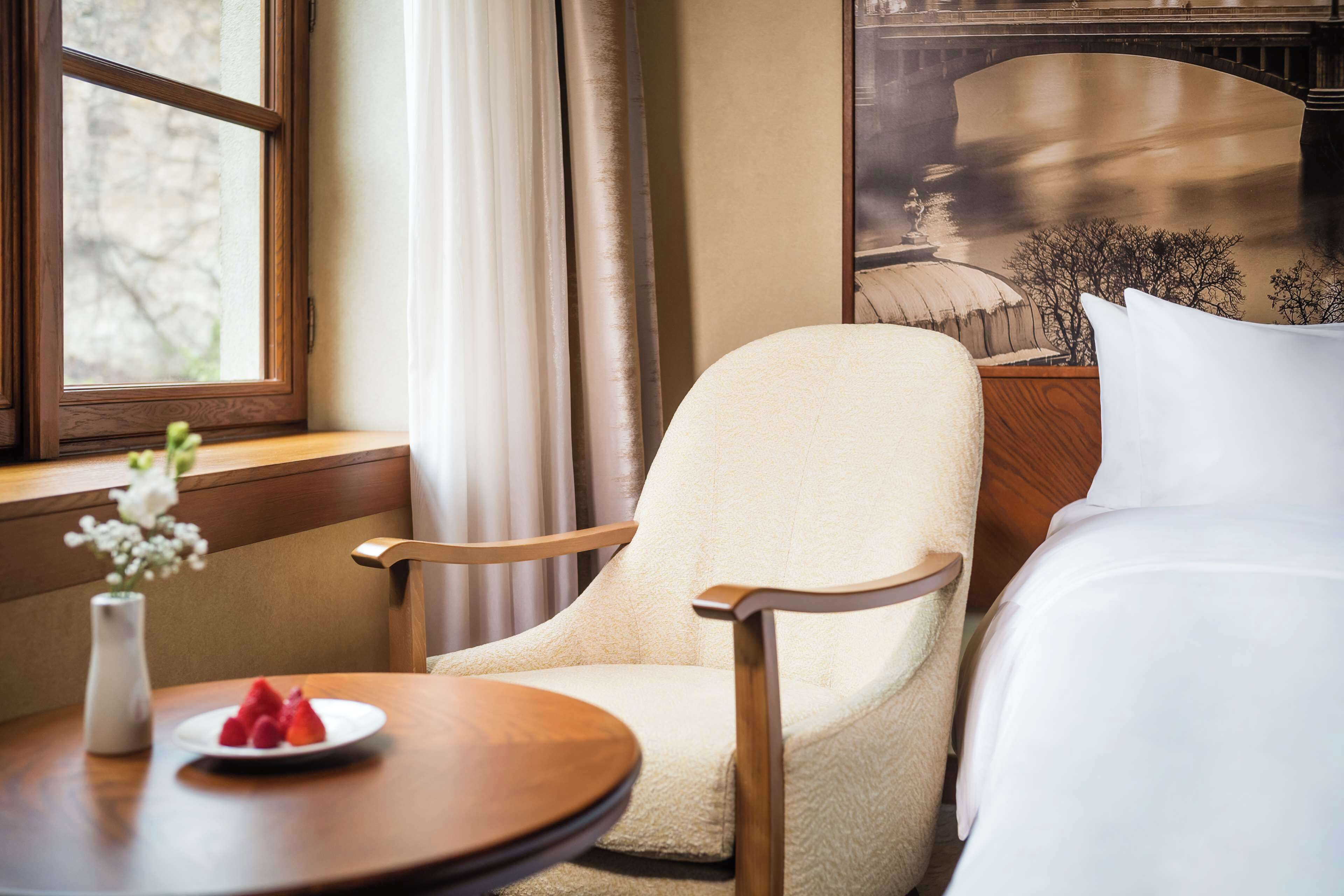 A hotel room with a bed, a chair, a round table with a plate of fruit, and a window with curtains.