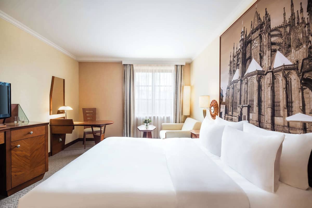 Habitación de hotel con una cama grande, un escritorio, una silla, un televisor y un cuadro de una catedral en la pared.
