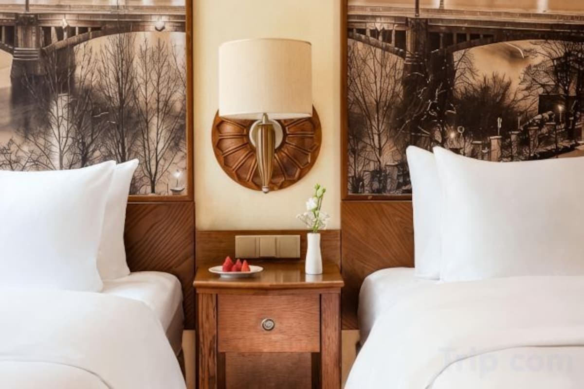 Une chambre d’hôtel avec deux lits, une table de chevet avec une lampe, un vase de fleurs et une assiette de fruits.