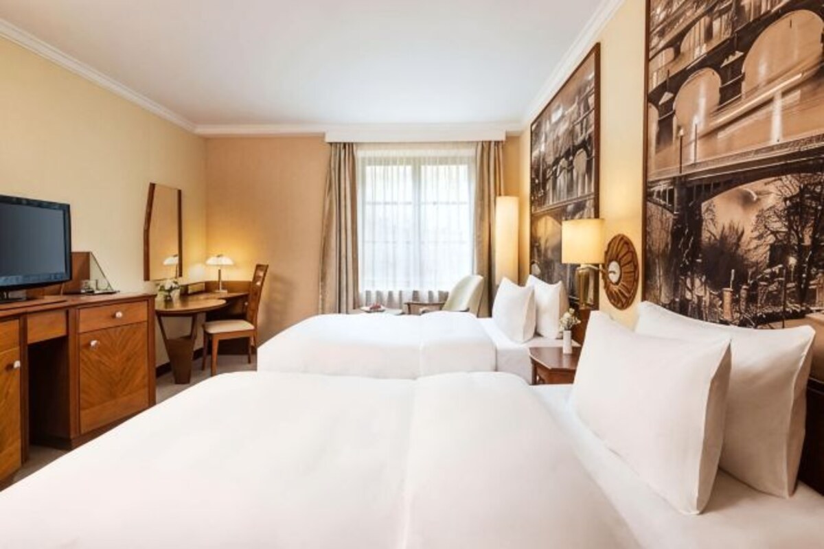 A hotel room with two beds, a desk, a television, and a large window with curtains.