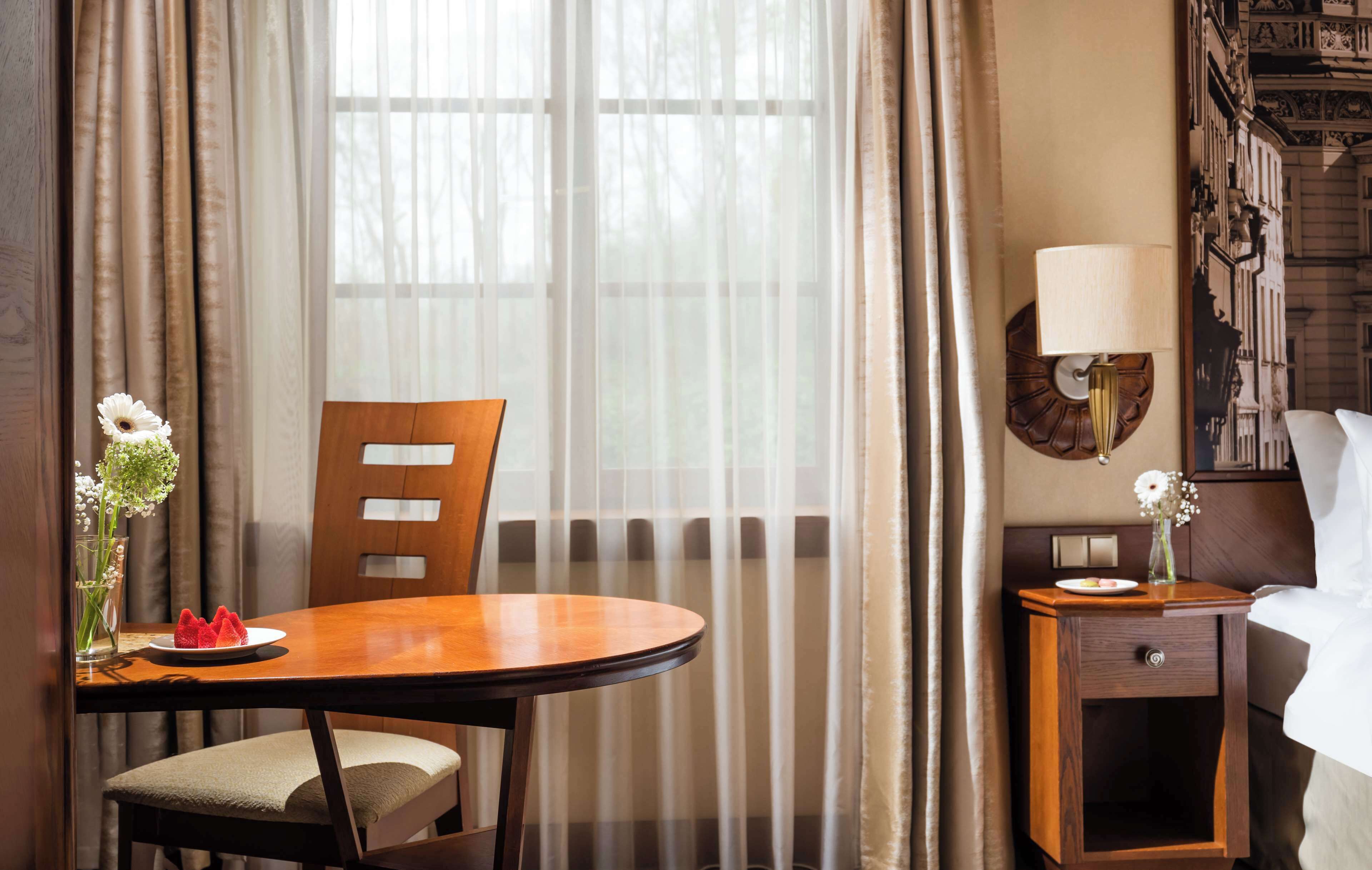 Habitación de hotel con una mesa de comedor de madera, una silla, una mesita auxiliar con una lámpara y una cama con ropa blanca de cama.