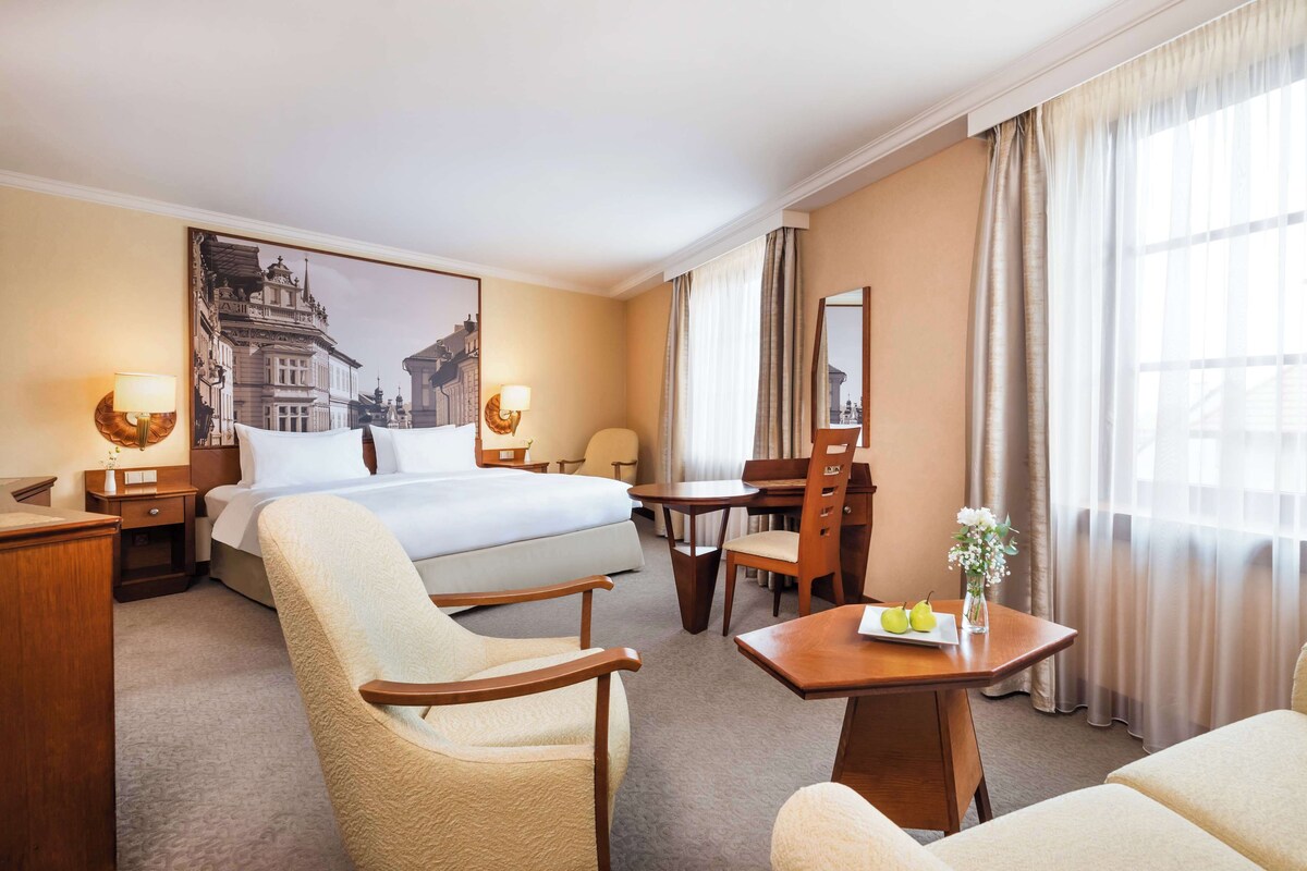 A hotel room with a large bed, a desk, two chairs, a small table with a vase of flowers, and a painting on the wall.