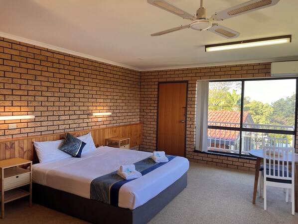 A hotel room with a brick wall, a wooden headboard, a bed with white and blue bedding, a nightstand, a desk with a chair, and a window with a view of buildings and trees. - Bayview Motor Inn (Eden)