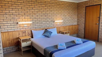 Una habitación de hotel con pared de ladrillo visto, cabecera de madera, cama con colcha azul y blanca, dos mesitas de noche con lámparas y una puerta de madera.