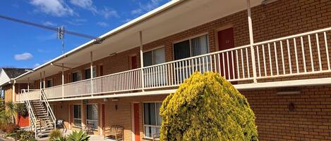 A two-story motel with a balcony on the second floor, a red door, and a well-maintained garden with various plants.