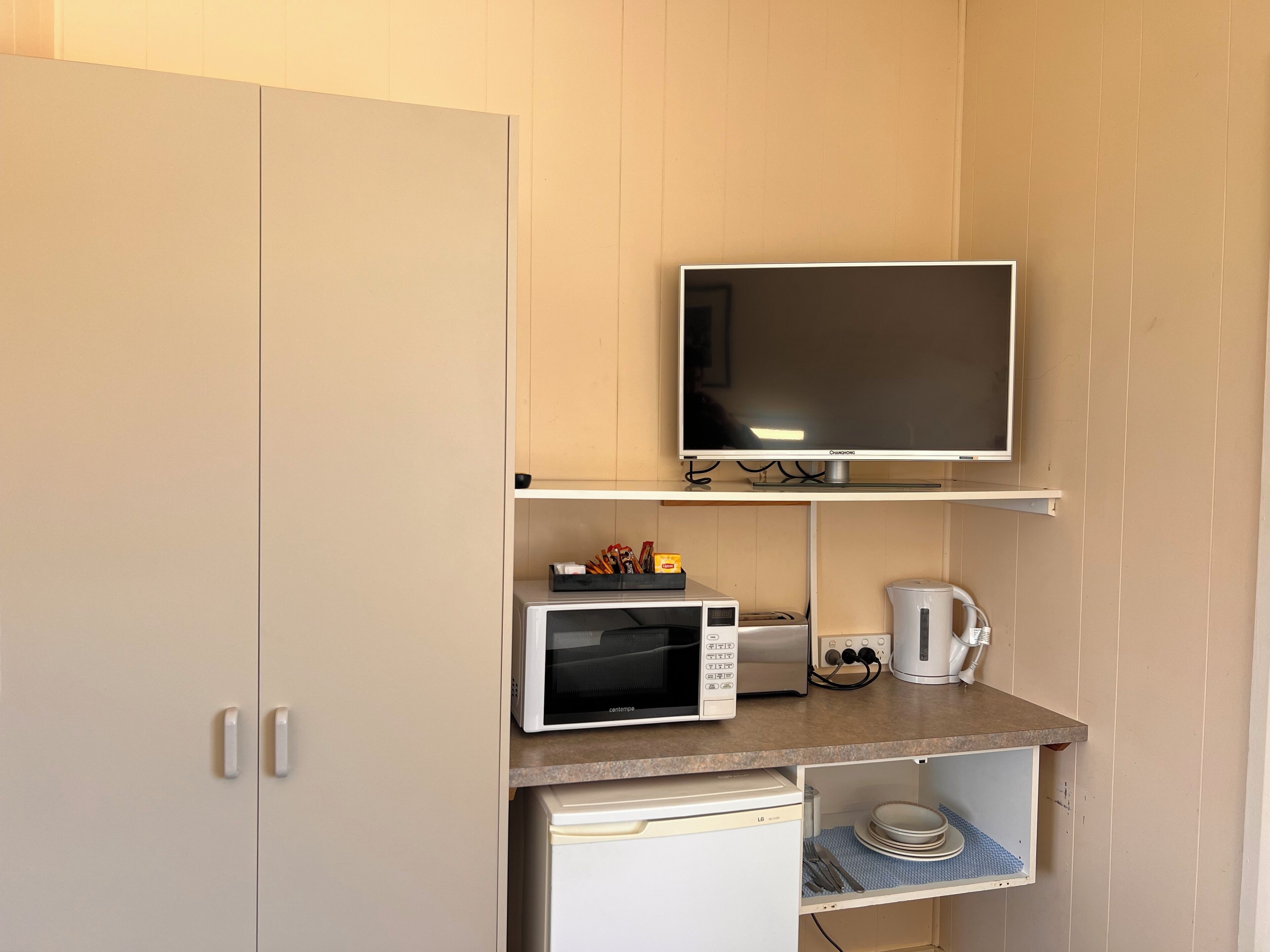 Une petite cuisine équipée d’un four à micro-ondes, d’un grille-pain et d’une machine à café, le tout installé sur un plan de travail, ainsi qu’un meuble sur lequel est posée une télévision.