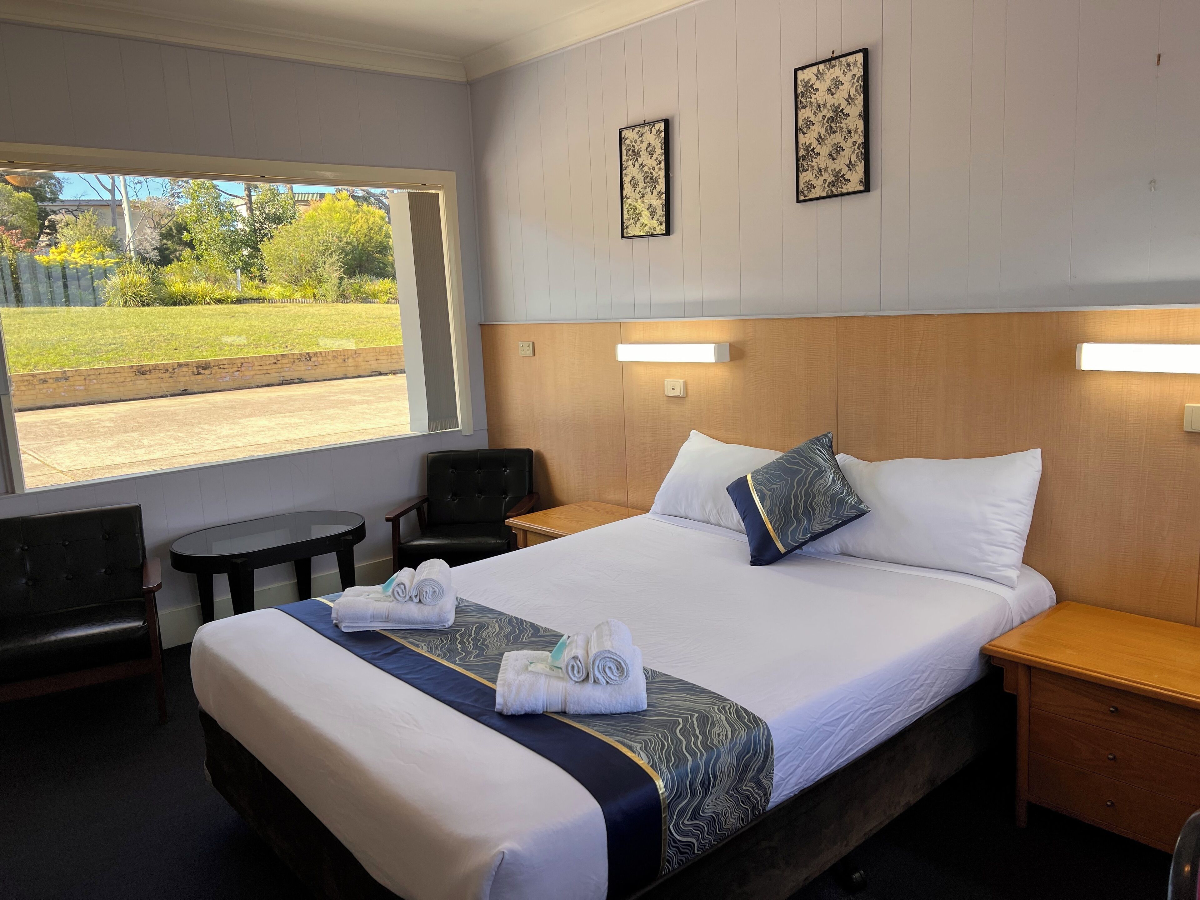 Habitación de hotel con una cama grande, dos mesitas de noche, una silla, una pequeña mesa y una ventana con vista a zonas verdes.