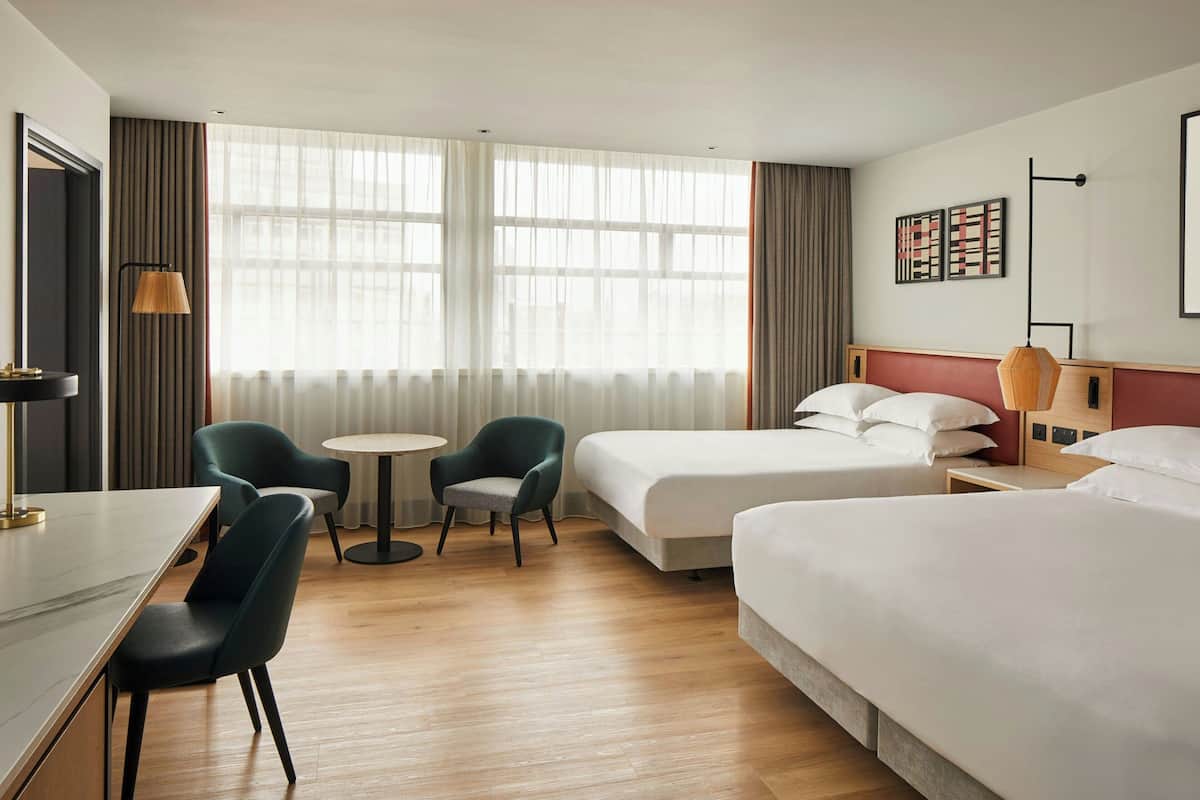 Ett hotellrum med två sängar, ett skrivbord, en stol, ett litet bord och en lampa.