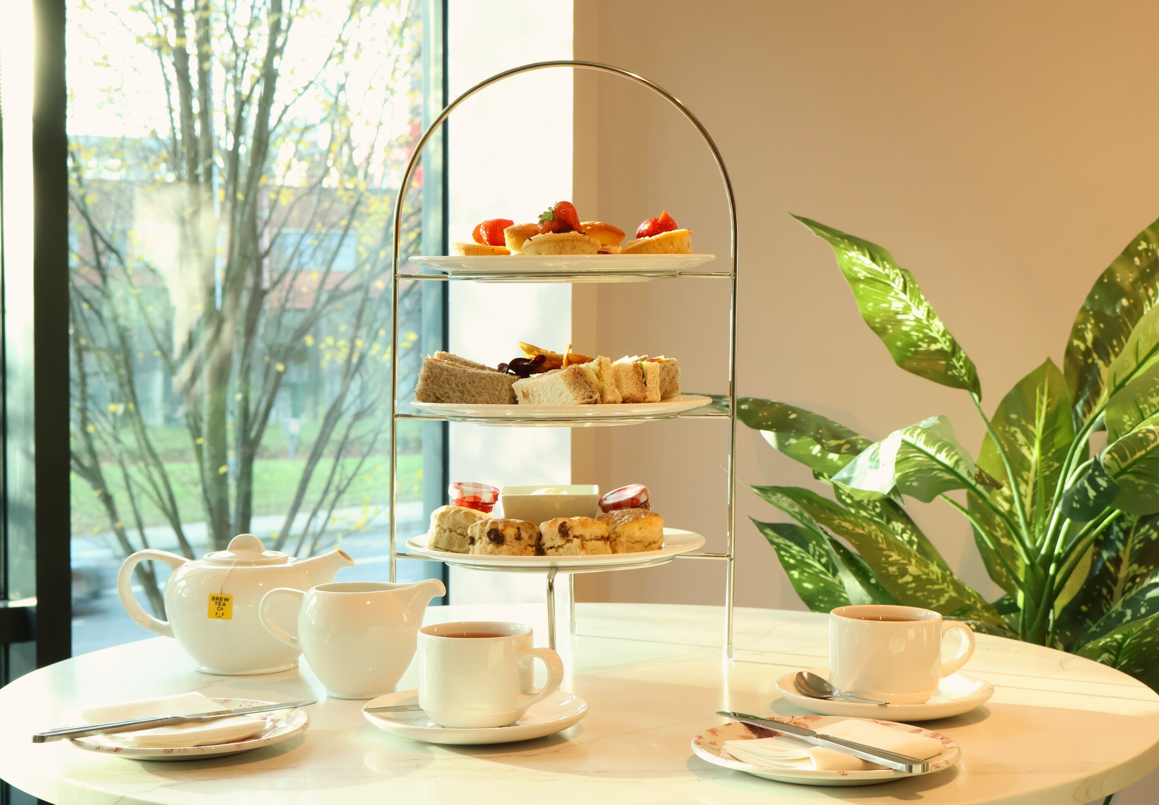 Un présentoir à trois niveaux, garni de diverses pâtisseries et de sandwichs, accompagné de théières, de tasses et de soucoupes, le tout disposé sur une table ronde.