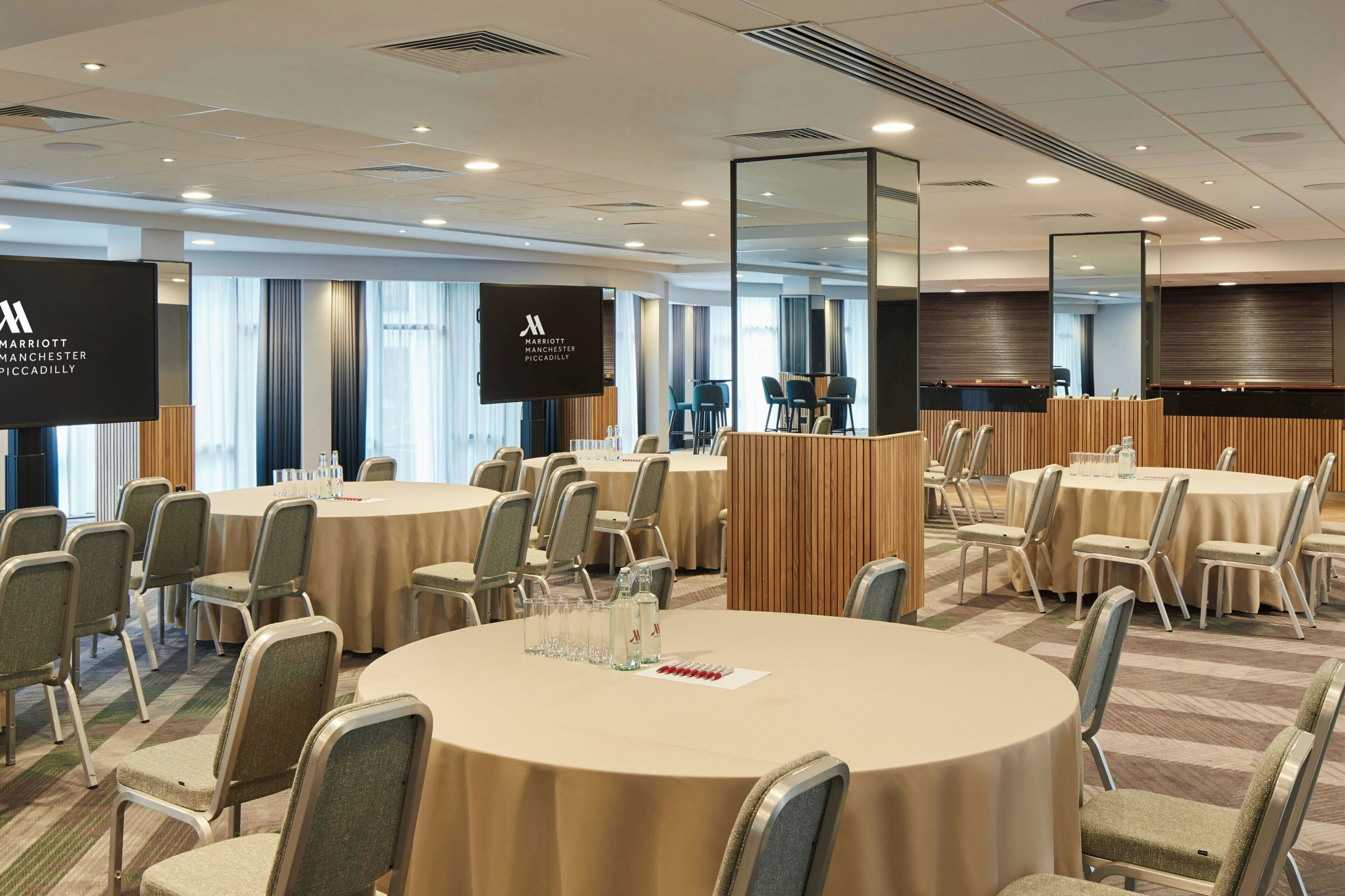 Un salón de conferencias con mesas redondas, sillas y una pantalla grande que muestra el nombre del hotel.