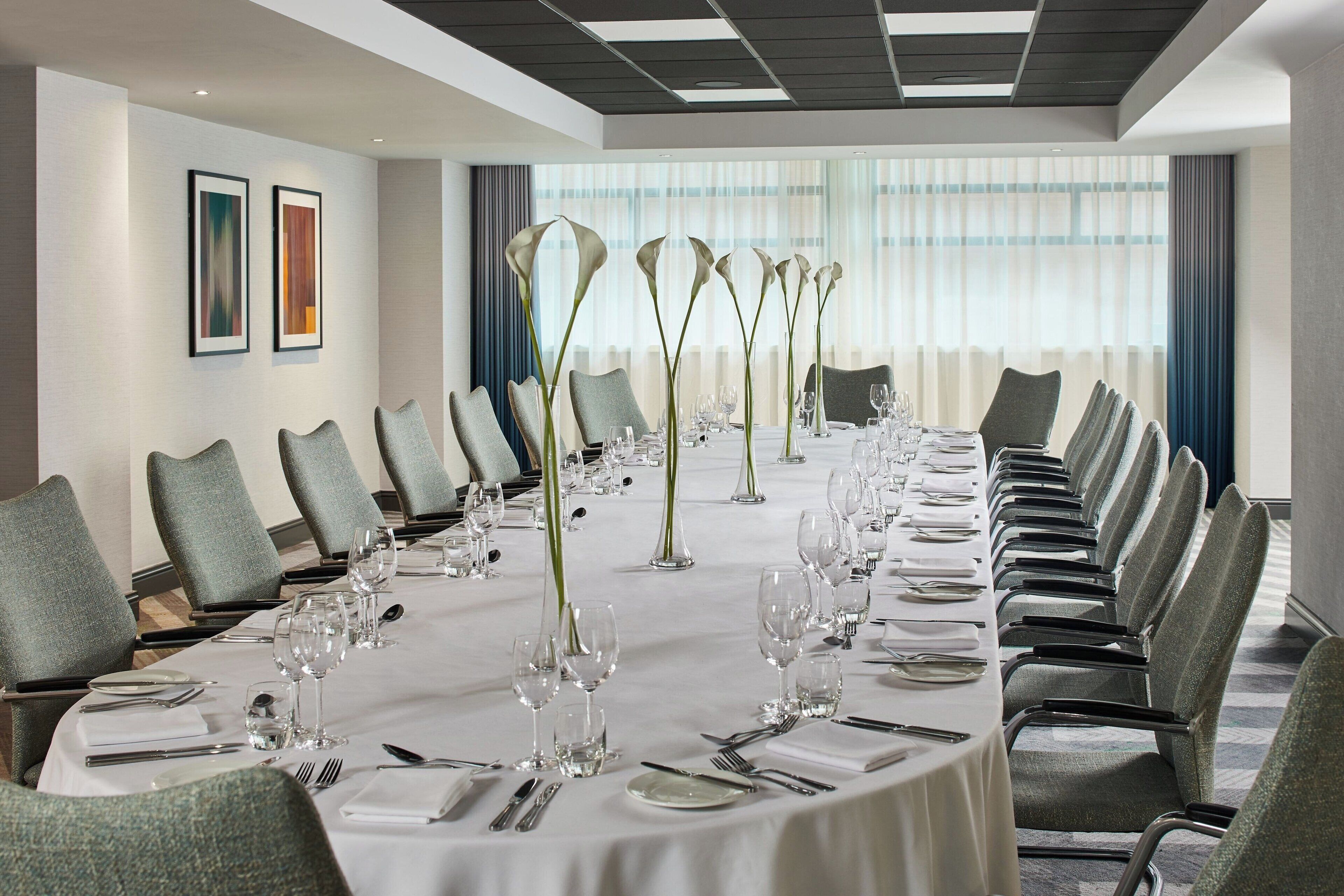 A conference room with a long table set for a meeting, surrounded by chairs, and adorned with tall vases.