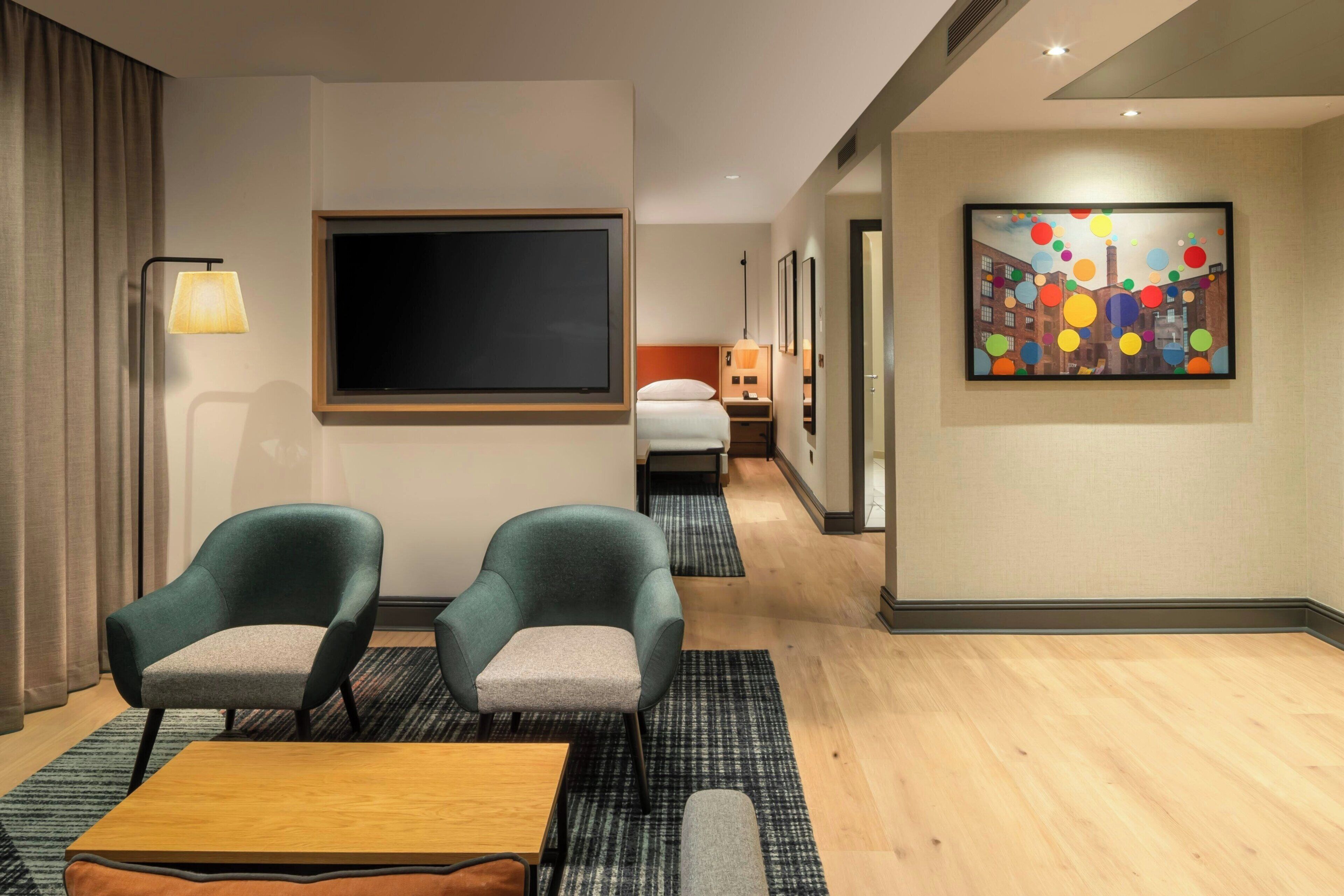 A hotel room with a TV, two armchairs, a wooden table, a bed, and a colorful artwork on the wall.