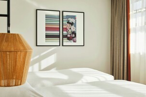 A hotel room with a bed, a wooden lamp, and two framed artworks on the wall.