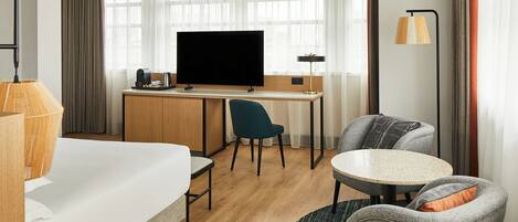 A modern hotel room with a bed, a desk, a television, a chair, and a round table.