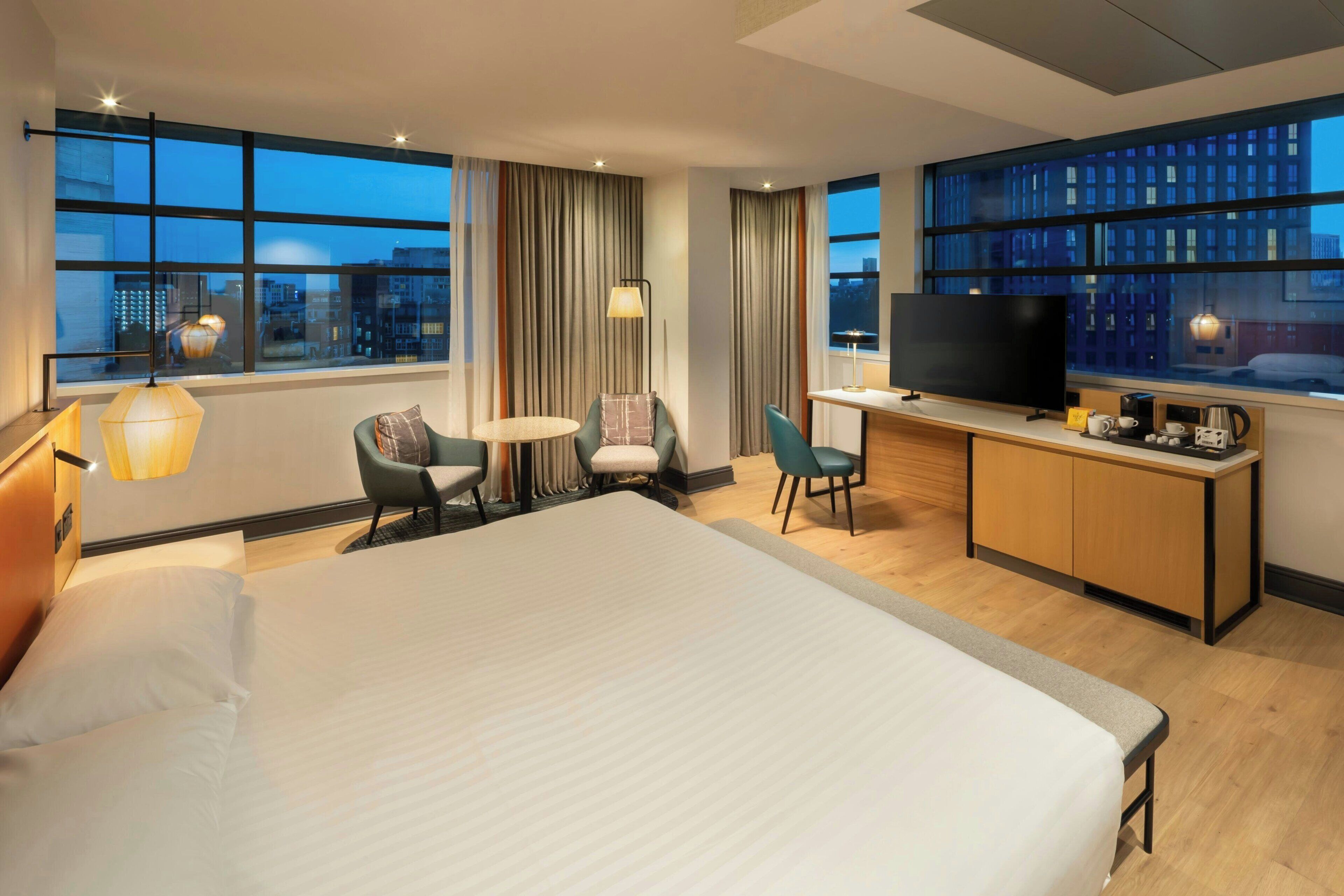 A hotel room with a large bed, a desk with a TV, a seating area with chairs, and large windows with a city view.