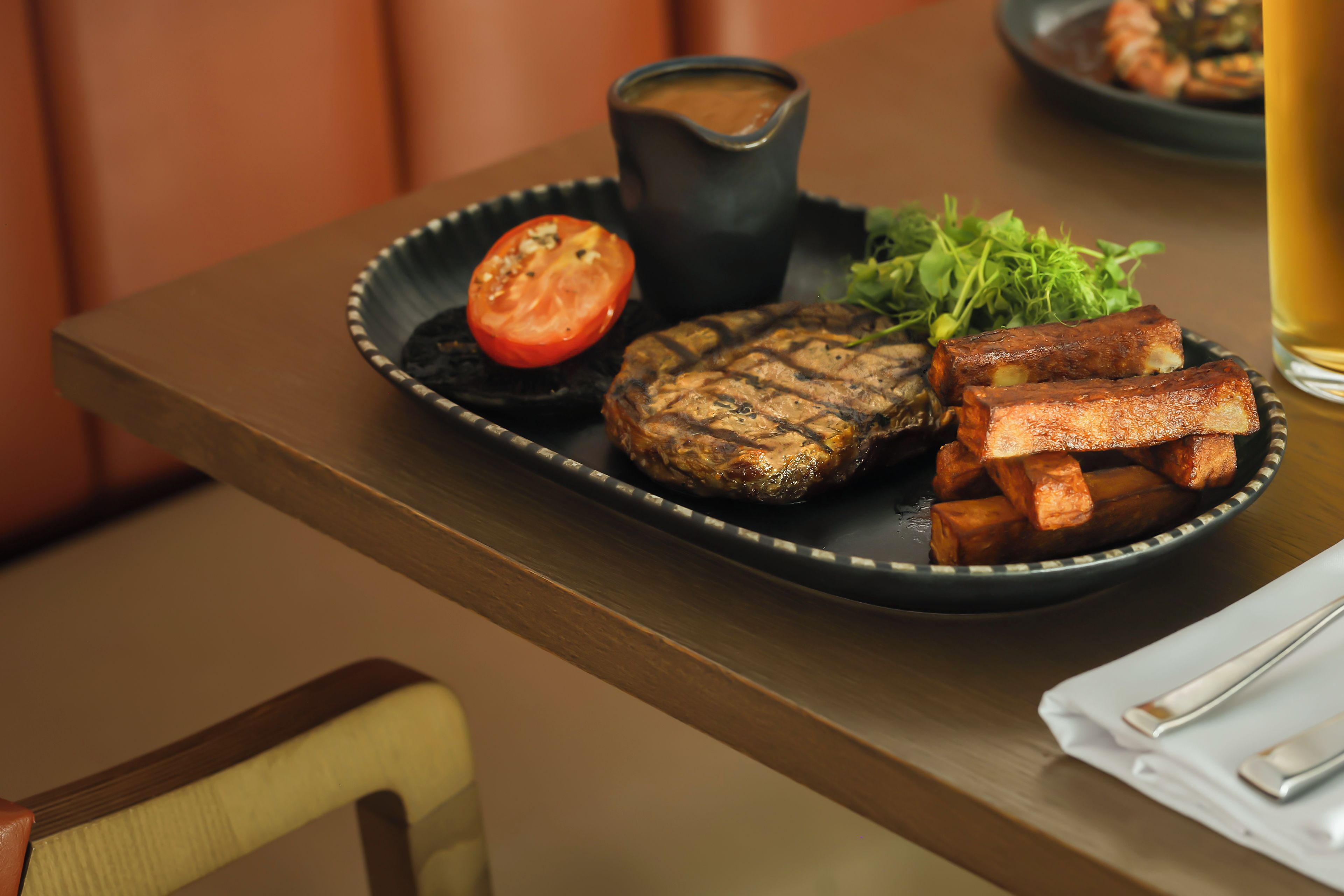 A plate with grilled meat, a grilled tomato, fries, and a small bowl of sauce, accompanied by a glass of beer.