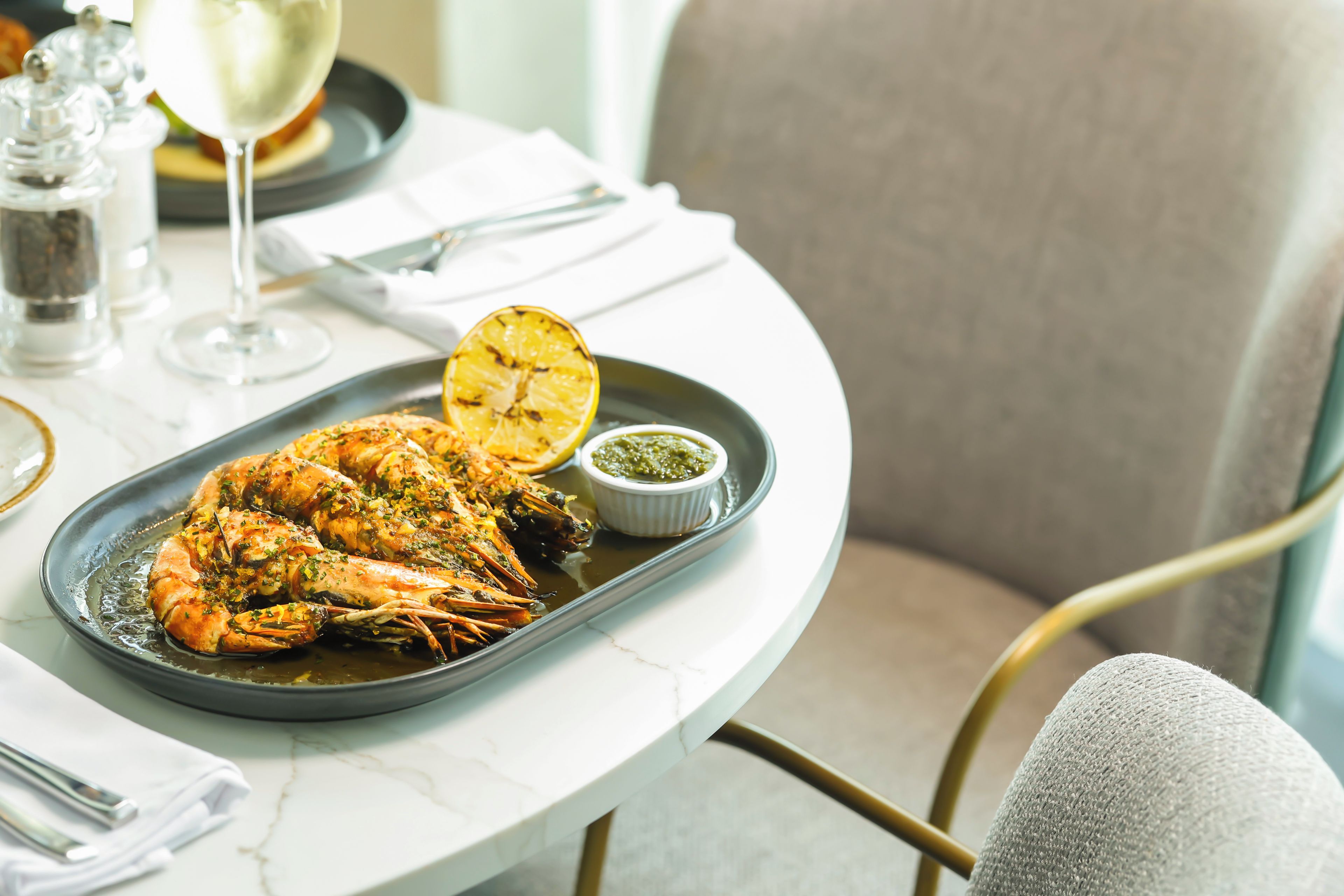 Un plat de fruits de mer garni d’une tranche de citron et nappé de sauce, présenté sur une assiette posée sur une table. On aperçoit également un verre de vin blanc et des salières et poivrières.