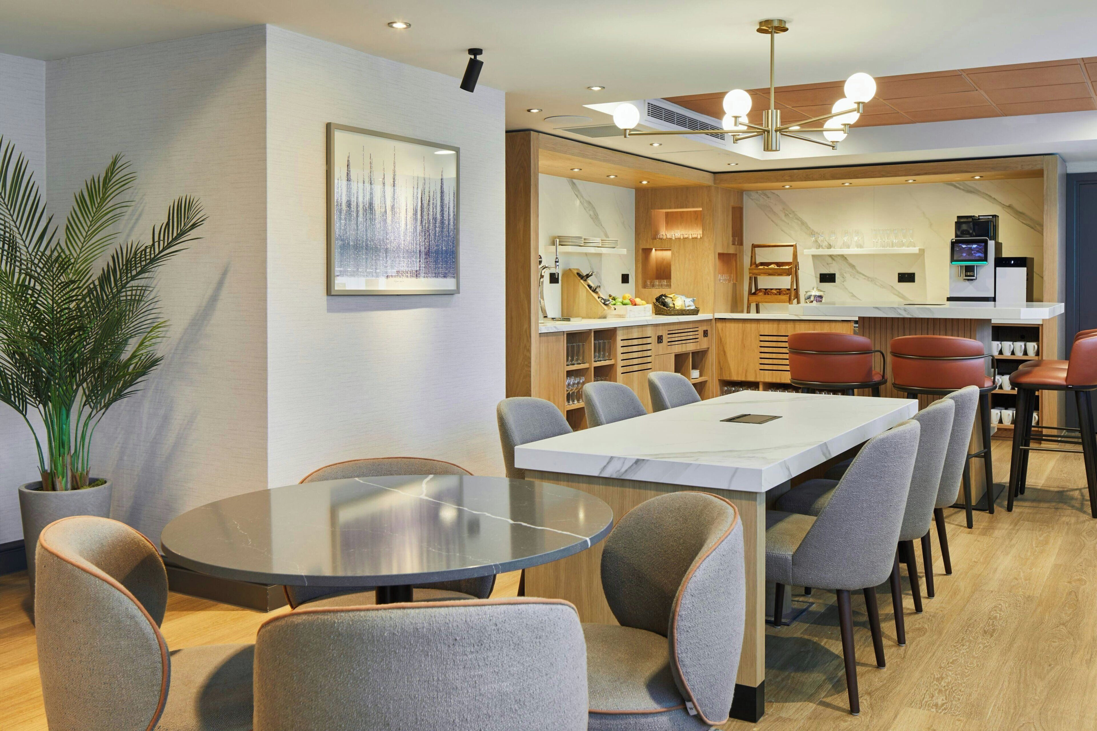 A modern dining area with a round table and grey upholstered chairs, a potted plant, and a kitchen area with a marble countertop and bar stools.