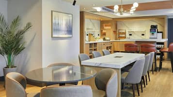 A modern dining area with a round table and grey upholstered chairs, a potted plant, and a kitchen area with a marble countertop and bar stools.