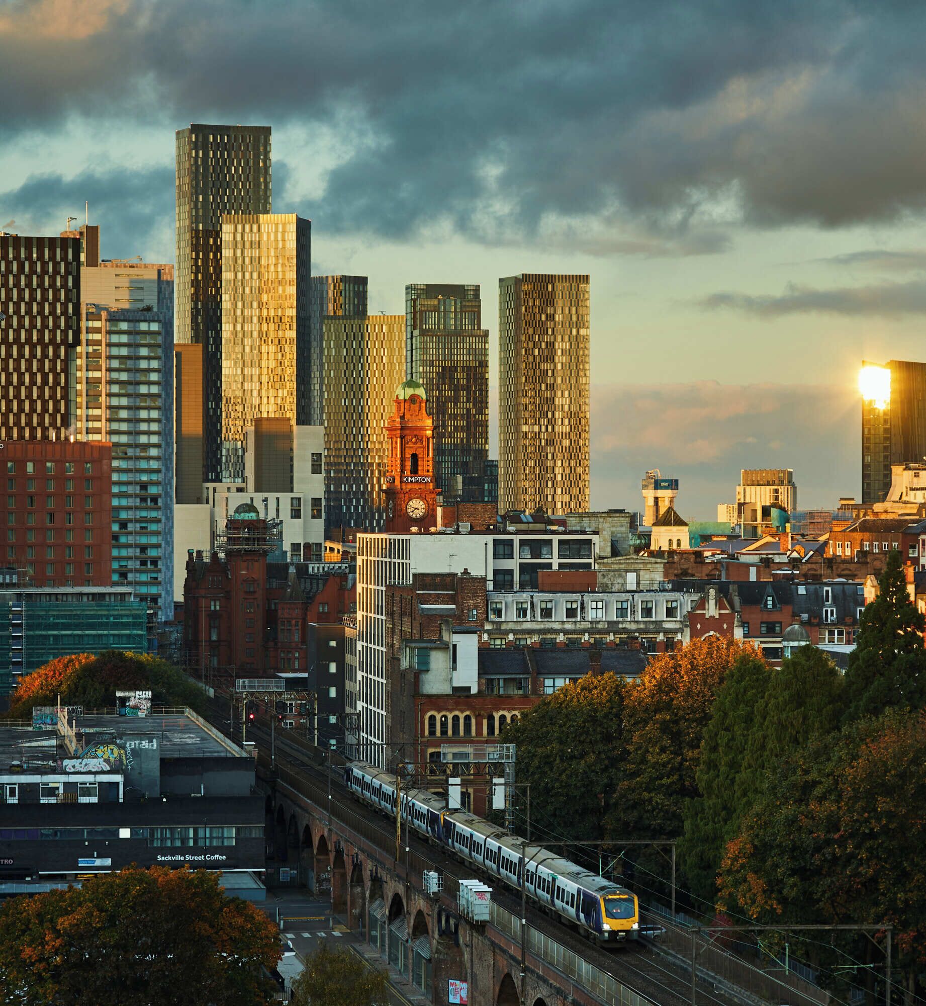A cityscape with modern high-rise buildings, a train on an elevated track, and a mix of older and newer architecture.