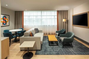 A modern hotel room with a sofa, chairs, a small table, a TV, and a large window with curtains.