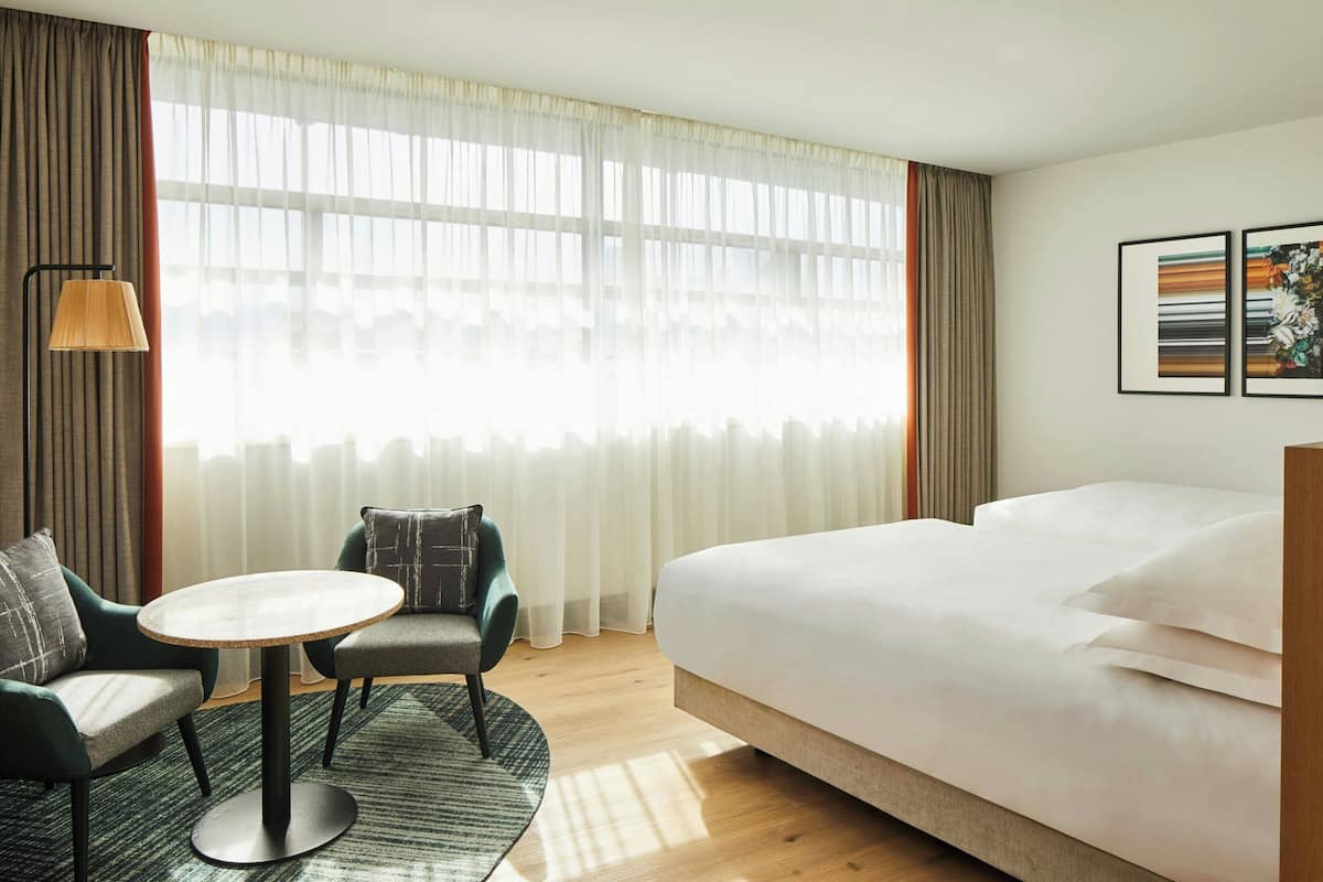 Ett hotellrum med en säng, två fåtöljer, ett litet runt bord och ett stort fönster med tunna, genomskinliga gardiner.