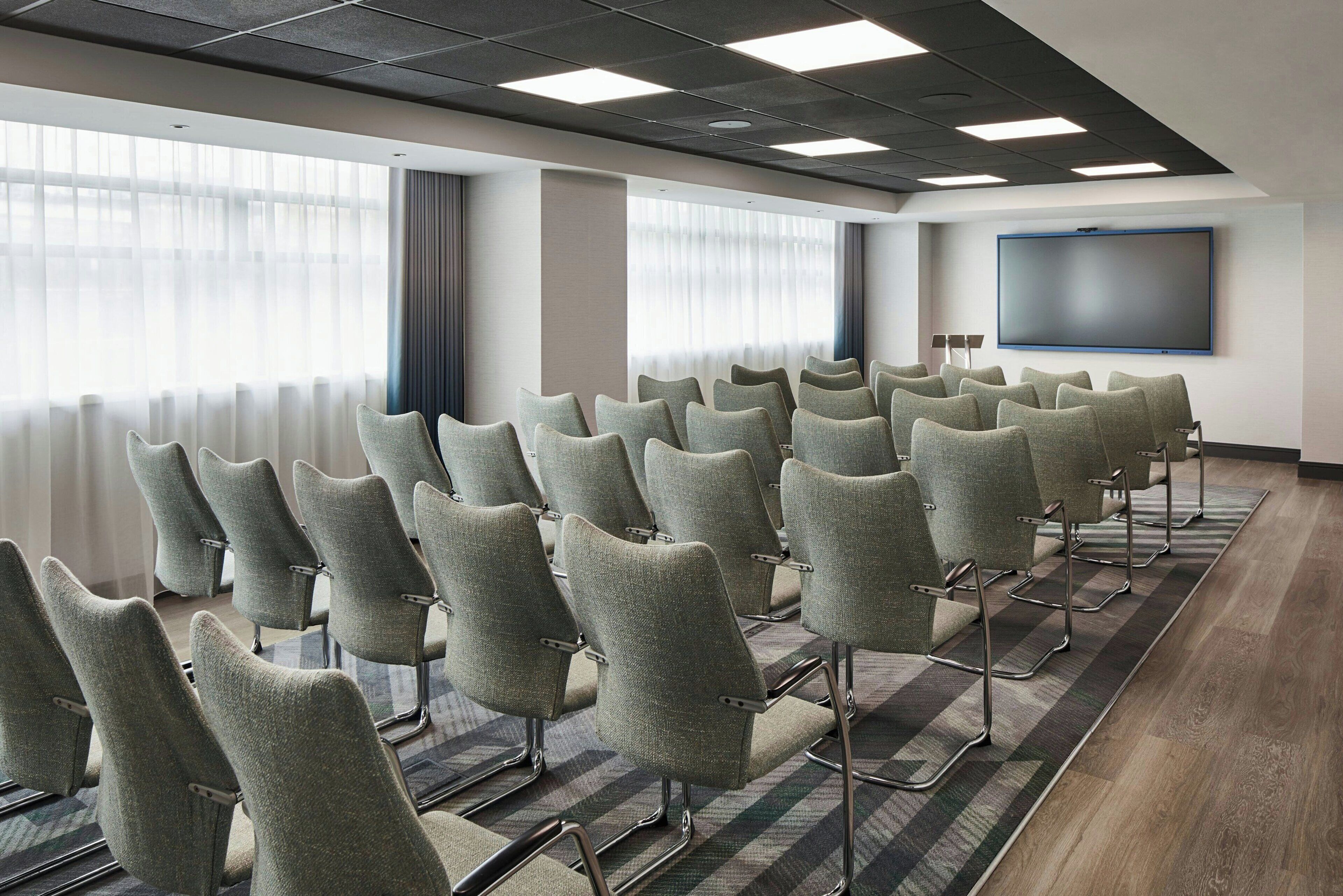 A conference room with rows of chairs, a large screen, and a window with curtains.