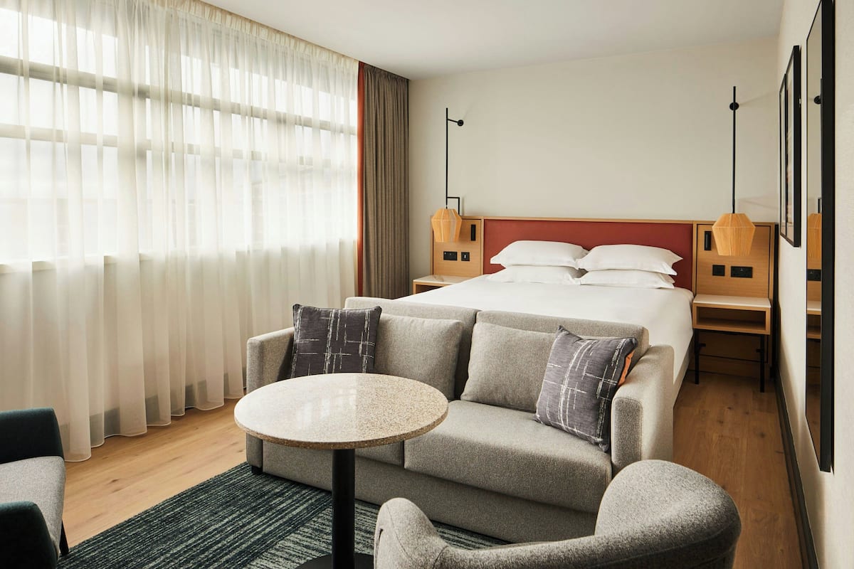 Ett hotellrum med en säng, en soffa, ett runt bord och en stol. Det finns två sängbord med lampor.